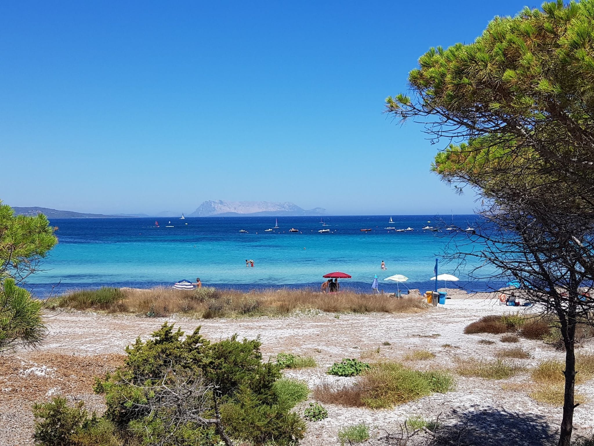 Een strand met helderblauw water, boten en mensen.