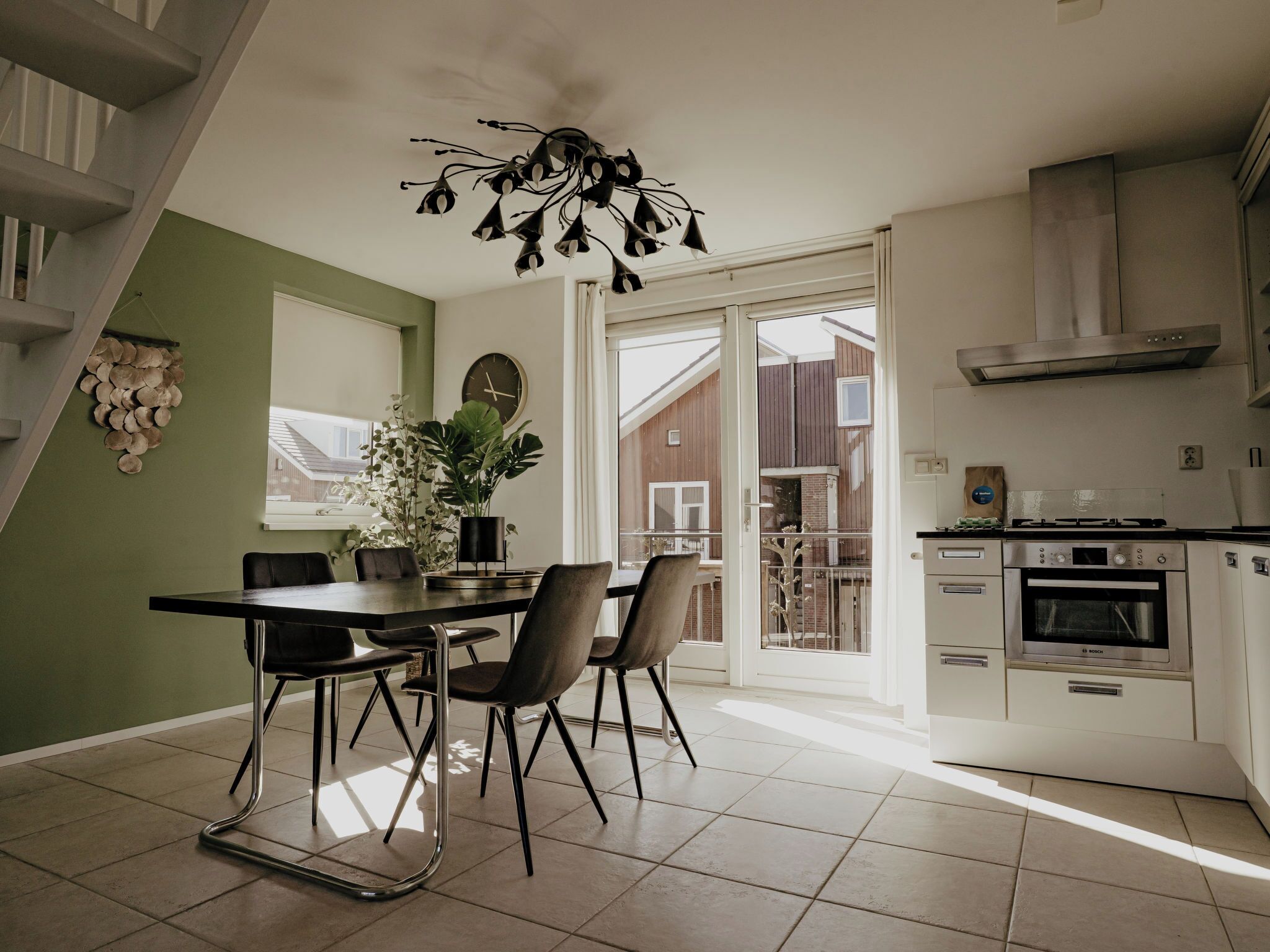 Een moderne keuken met een inbouwoven, een eethoek met een tafel en stoelen, en een uitzicht op het buitengebeuren door grote ramen.