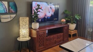 A living room with a flat-screen TV on a wooden cabinet, two potted plants, a floor lamp, and a round mirror on the wall. - Ace on Par - Central Dunsborough (Dunsborough)