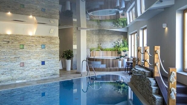 En indendørs swimmingpool med en stenvæg, en trætrap og et barområde med planter.