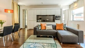 A modern living room with a glass coffee table, a grey sofa with orange cushions, a dining area with black chairs, and a kitchen with white cabinets and a black refrigerator.