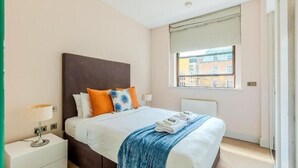 A neatly made bed with a blue throw blanket, a white nightstand with a lamp, and a window with a view of buildings.