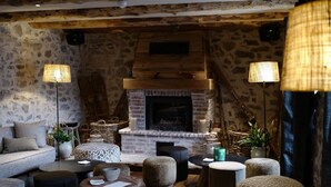 Ein gemütliches, rustikales Wohnzimmer mit Steinwand, Holzbalken, Kamin und verschiedenen Sitzgelegenheiten.