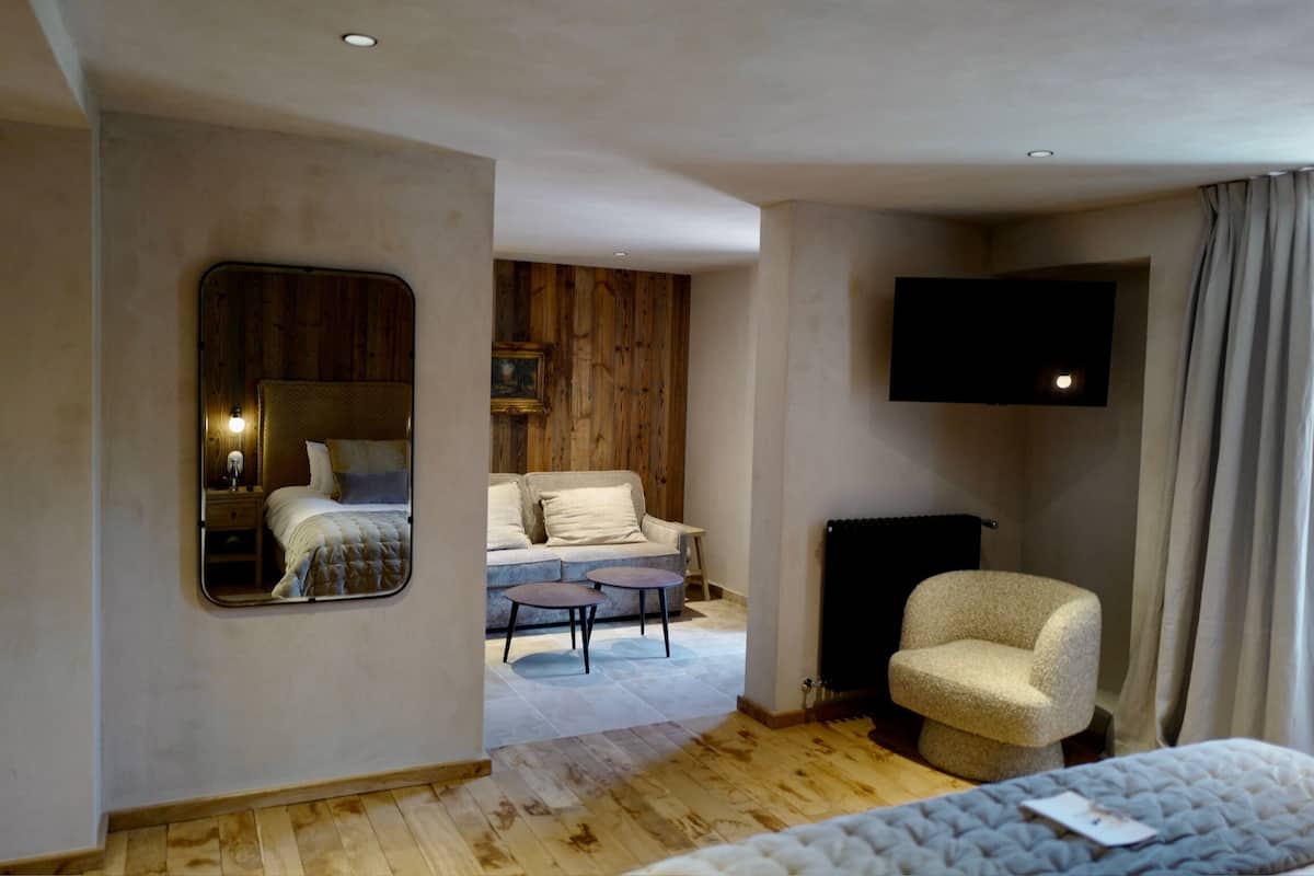 A hotel room with a wooden floor, a bed with a white duvet, a sofa, a small round table, a chair, a flat-screen TV mounted on the wall, and a window with curtains.