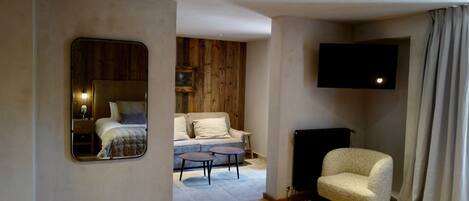 A hotel room with a wooden floor, a bed with a white duvet, a sofa, a small round table, a chair, a flat-screen TV mounted on the wall, and a window with curtains.