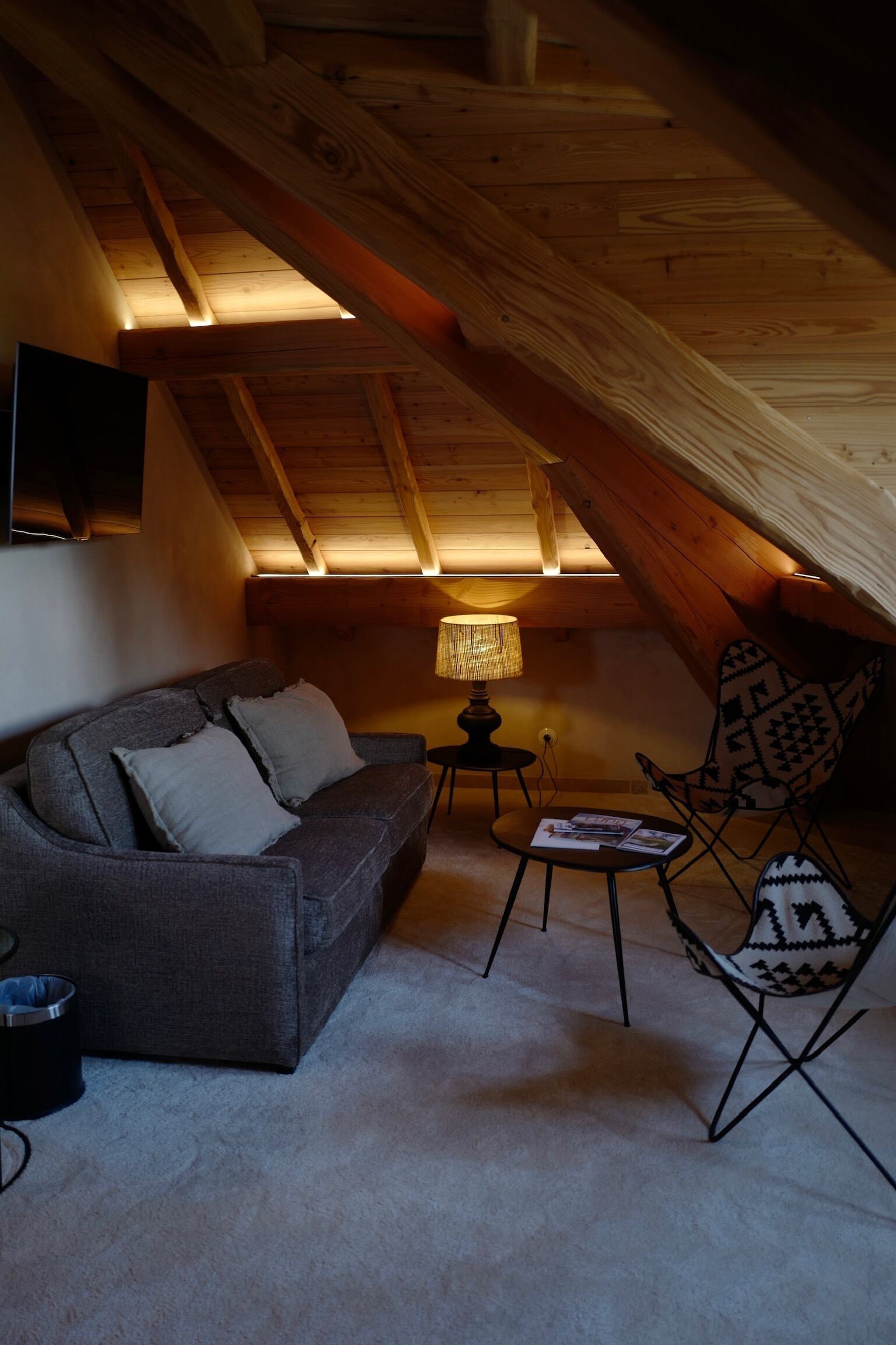 Un dormitorio en el ático con techo inclinado, un sofá, una silla estampada, una mesita con una lámpara y un televisor montado en la pared.