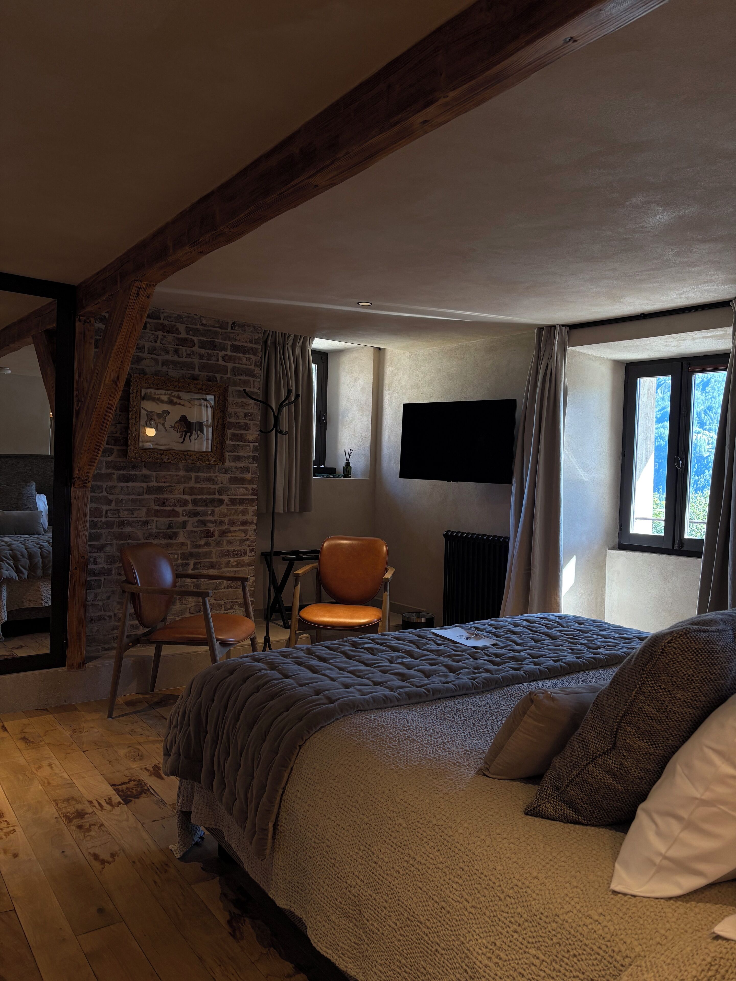 A cozy bedroom with exposed wooden beams, a brick wall, a large bed, a chair, and a TV mounted on the wall.