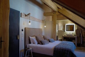 Une chambre avec un plafond en bois, un lit recouvert de plusieurs oreillers, une table de chevet et un petit tabouret.