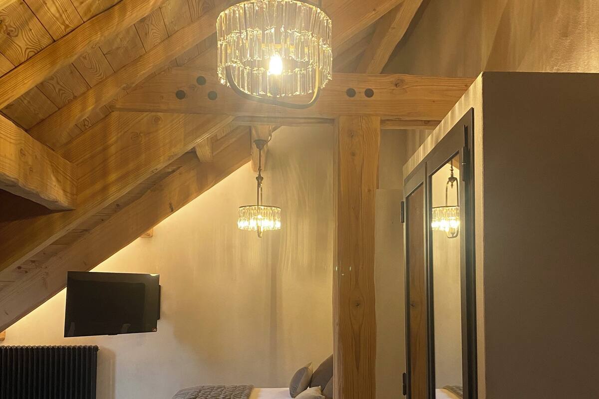 A bedroom with a sloped ceiling, wooden beams, a bed with a grey quilt, a bedside table, a small rug, and a chandelier.