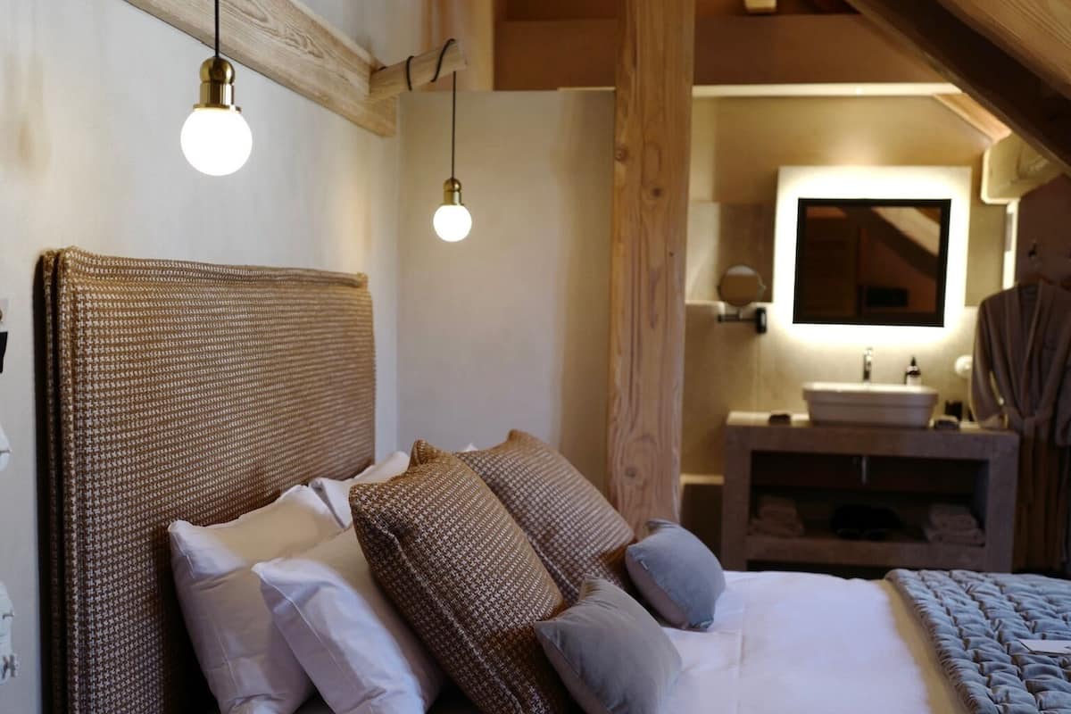 A bedroom with a large bed, wooden beams, a small stool, and a bathroom with a mirror and sink.