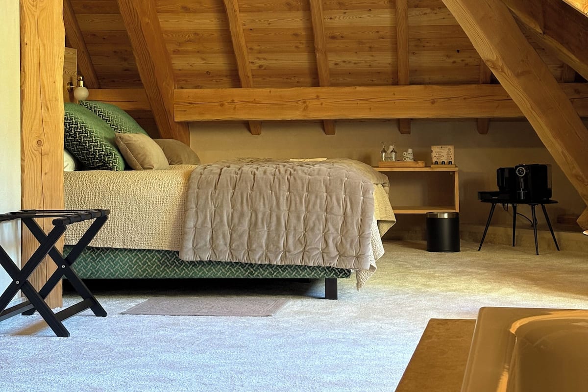 A cozy attic bedroom with a wooden ceiling, a bed with a green and white bedspread, and a small table with a kettle.