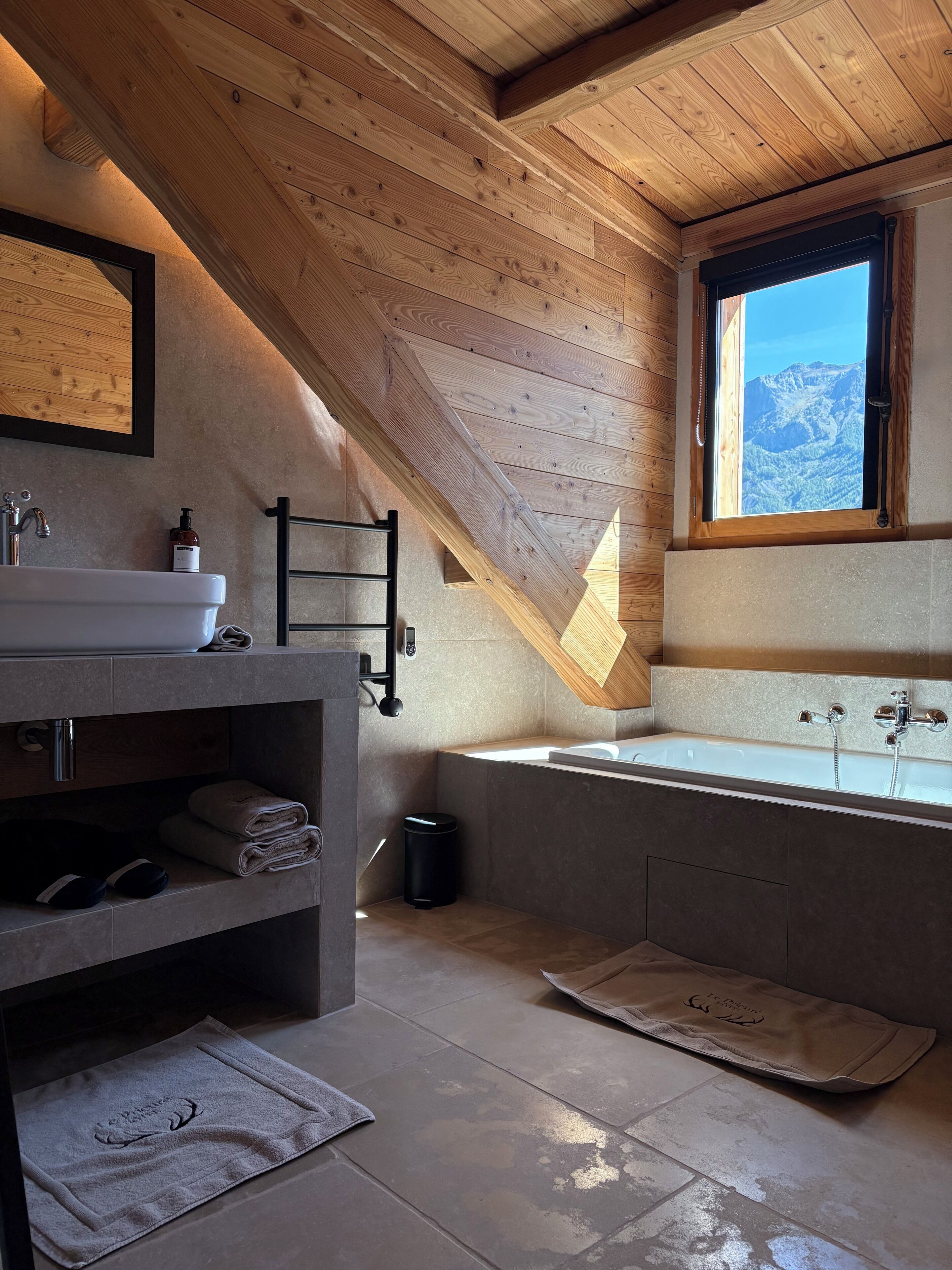 A modern bathroom with a wooden ceiling, a large window with a mountain view, a bathtub, and a sink with a towel rack.