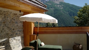 Eine Terrasse mit Steinmauer, Holzbalken, einem grünen Sofa, einem weißen Sonnenschirm und Blick auf die Berge.