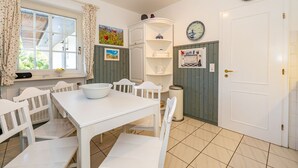 A dining area with a white table and chairs, a window with curtains, a wall-mounted electrical panel, and a door with a light switch. - Rüm hart - Spacious vacation apartment Rüm hart on Sylt with terrace for max. 5 persons. (Sylt-Ost)