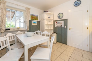 A dining area with a white table and chairs, a window with curtains, a wall-mounted electrical panel, and a door with a light switch.