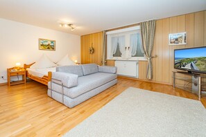 Ein gemütliches Wohnzimmer mit grauem Sofa, Holzboden, Fernseher und einem Fenster mit Vorhängen.
