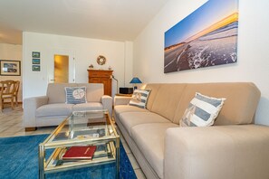 A living room with a beige sofa, a glass coffee table, and a blue rug. There is a painting on the wall and a wooden cabinet.