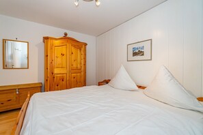 Un dormitorio con un armario de madera, una cama con ropa de cama blanca, una mesita de noche y un cuadro enmarcado en la pared.
