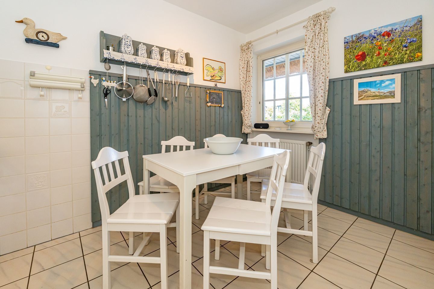 A kitchen with white cabinets, a white table, and chairs. There are hanging pots and pans, a window with curtains, and a painting on the wall.
