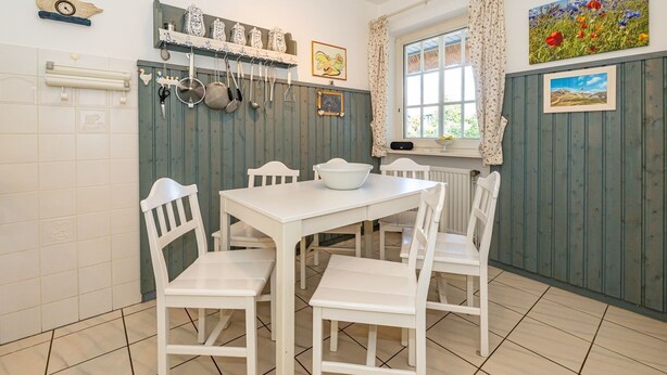Une cuisine avec des meubles blancs, une table blanche et des chaises. On y voit des casseroles et des poêles suspendues, une fenêtre avec des rideaux et un tableau accroché au mur.