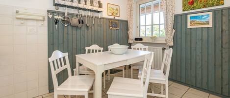 A kitchen with white cabinets, a white table, and chairs. There are hanging pots and pans, a window with curtains, and a painting on the wall.