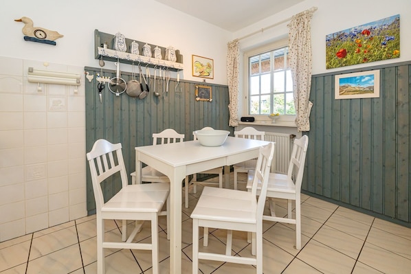 Una cocina con armarios blancos, una mesa blanca y sillas. Hay ollas y sartenes colgadas, una ventana con cortinas y un cuadro en la pared.