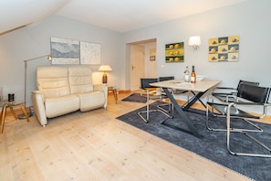 A modern living room with a beige sofa, a dining table with four chairs, and artwork on the walls. - Goofys Will - Modern vacation apartment Goofys Will on Sylt with comfort and (Sylt)