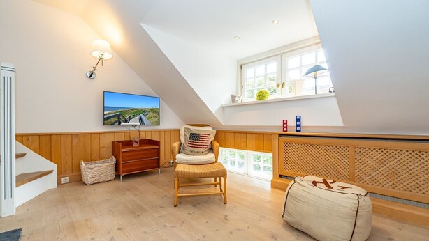 A loft bedroom with a sloped ceiling, wooden flooring, a wooden dresser, a wicker chair, a bean bag, a TV on the wall, and a window with a view of greenery. - Goofys Will - Modern vacation apartment Goofys Will on Sylt with comfort and (Sylt)