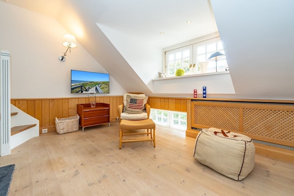 Une chambre à coucher aménagée dans un espace mansardé, avec un plafond en pente, un plancher en bois, une commode en bois, un fauteuil en osier, un pouf, un téléviseur fixé au mur et une fenêtre donnant sur un paysage verdoyant.