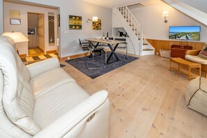 A modern living room with a white leather sofa, a dining area with a table and chairs, a staircase, and a television displaying a scenic view.