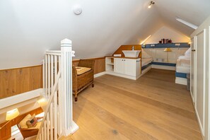 A loft bedroom with a wooden floor, a white staircase, a wicker chair, a white cabinet with a sink, and a bed with a blue headboard.