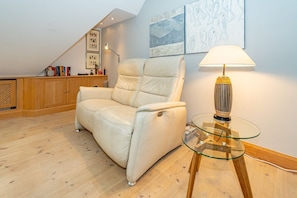 A modern living room with a leather armchair, a glass coffee table, and a lamp. There is a wooden cabinet and a loft bed with a sloped ceiling. - Goofys Will - Modern vacation apartment Goofys Will on Sylt with comfort and (Sylt)