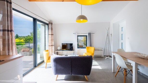 Un salon moderne avec un canapé, une table à manger et un téléviseur. Une grande fenêtre offre une vue sur l’extérieur.