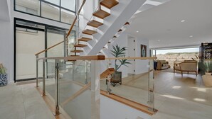 A modern interior with a glass staircase, a living area with a sofa and coffee table, and a view of the outdoors. - Time and Tides - Beachfront, Stunning Ocean Views. (THE ENTRANCE NORTH)