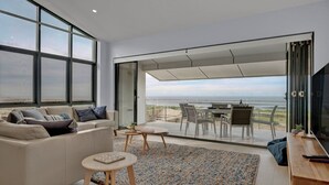 A modern living room with a large window, a sofa, a coffee table, and a dining area with chairs and a table. - Time and Tides - Beachfront, Stunning Ocean Views. (THE ENTRANCE NORTH)