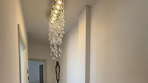 Un couloir étroit, orné d’un lustre, d’un radiateur et d’une chaise en bois.