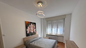 Une chambre moderne avec un plancher en bois, un lit recouvert d’une couette matelassée, une table de chevet, un radiateur et une fenêtre agrémentée de rideaux à motifs.