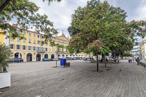 A public square with a large tree, a yellow building with arches, and a blue trash bin.