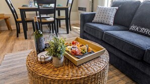 Un salon avec un canapé gris, une table basse en osier sur laquelle se trouve une bouteille de vin, une plante et un plateau de collations.