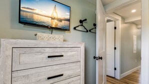 Une pièce comprenant une commode en bois, un téléviseur fixé au mur qui diffuse une image d’un voilier, et une porte donnant sur une autre pièce.