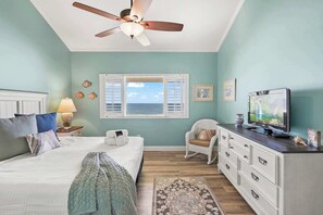 Un dormitorio con cama, ventilador de techo, televisor, cómoda y ventana con contraventanas.