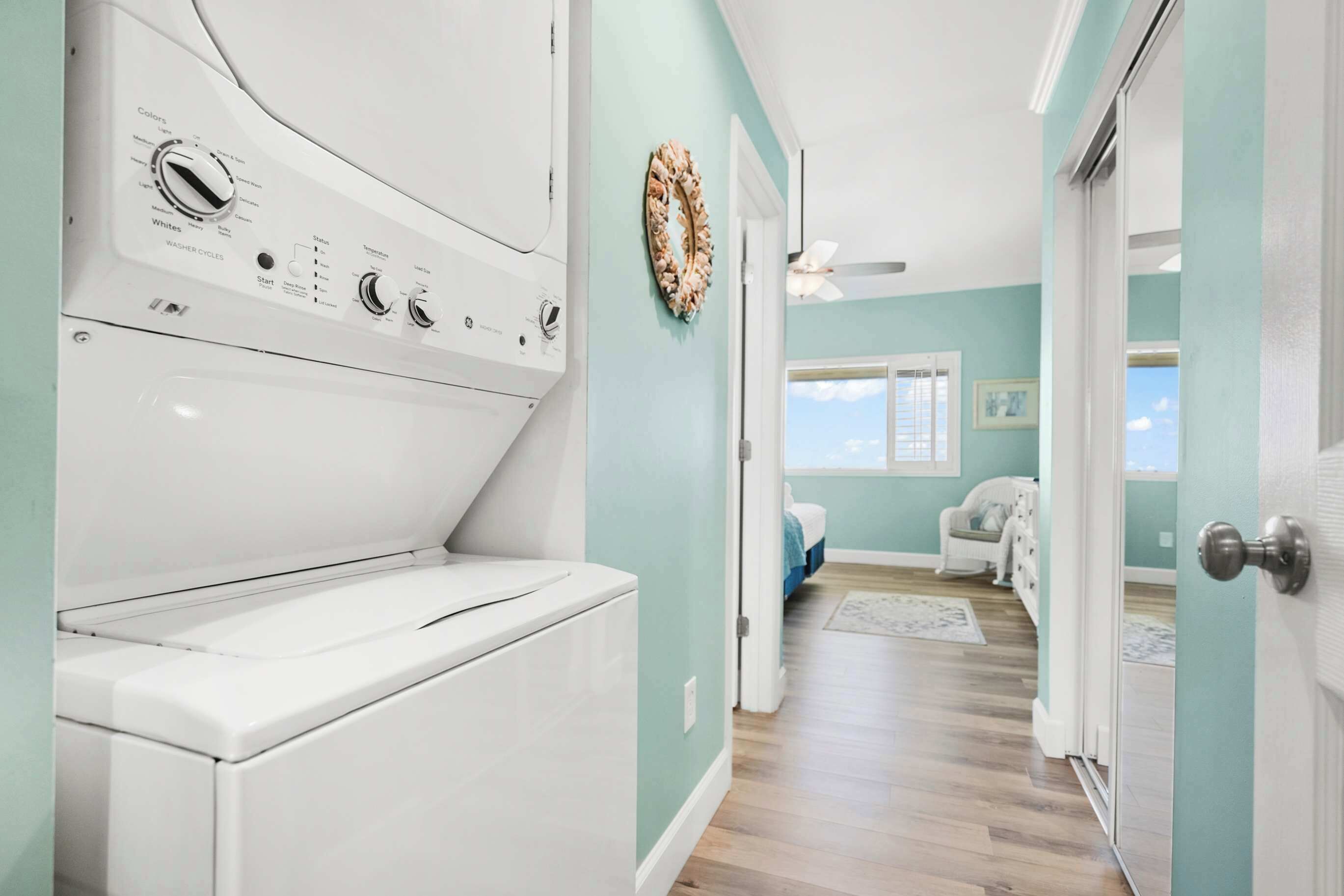 Un lavadero con lavadora y secadora de carga frontal, una puerta que da a un dormitorio con cama y sillón, y vista al cielo a través de una ventana.