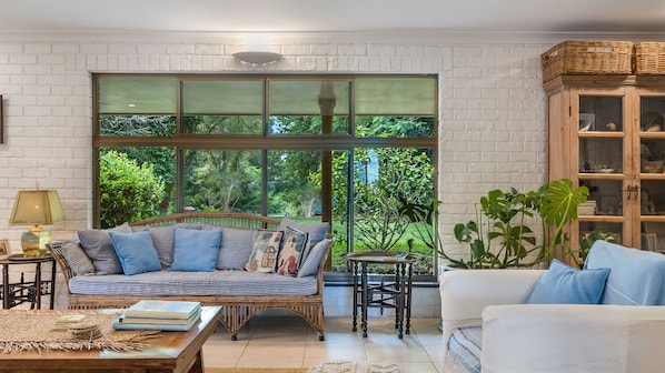 A cozy living room with a large window, a wicker sofa, a white armchair, and a wooden cabinet.