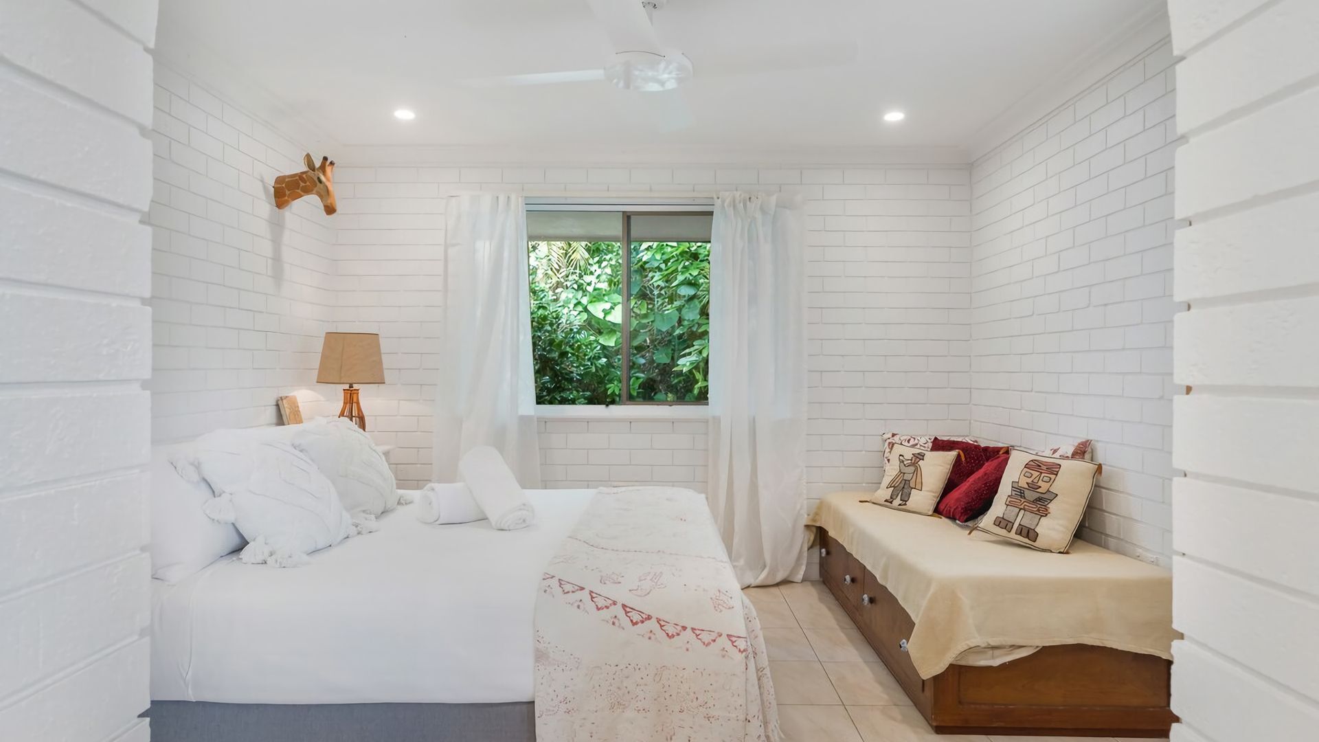 A bedroom with a bed, a bench, a lamp, and a window with a view of greenery.
