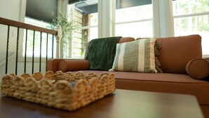 Una sala de estar acogedora con un sofá marrón, una cesta de mimbre y vistas a la vegetación a través de amplios ventanales.