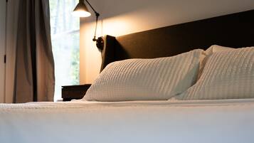 Una cama bien hecha con ropa blanca, cabecera oscura y lámpara de noche.