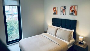 A modern bedroom with a large window, a bed with a dark headboard, and three abstract wall art pieces.