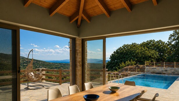 A dining area with a wooden table and chairs, a hanging chair, and a view of a swimming pool and landscape. - Bellevia Stone Pool Villa (Nea Styra)