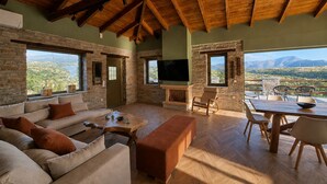 A spacious living area with a stone fireplace, a sofa, a television, and a dining table with chairs. - Bellevia Stone Pool Villa (Nea Styra)