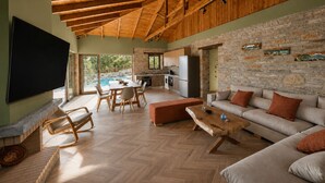 A spacious living area with a stone wall, wooden ceiling, and a pool visible through large windows. - Bellevia Stone Pool Villa (Nea Styra)